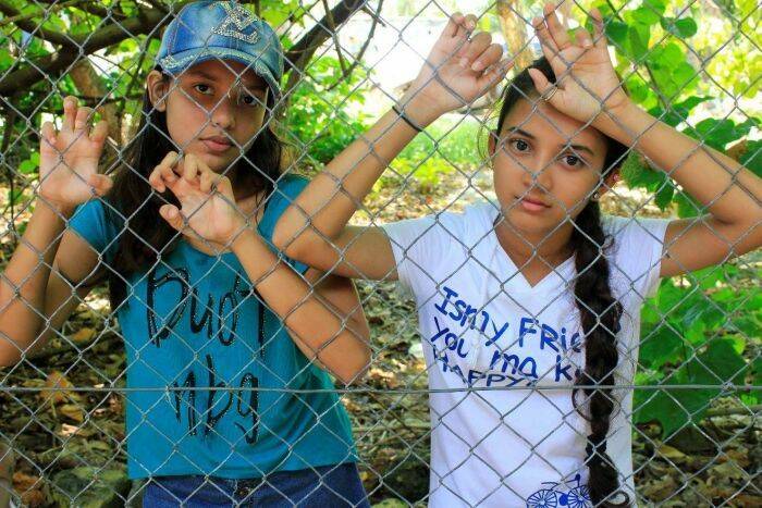 Two young refugee girls stand behind a barbed wire fence in Nauru. 
Image via ABC News.